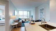 This single-floor residence is designed with a contemporary aesthetic, combining clean lines, neutral tones, and high-quality finishes throughout. The open-concept living area is anchored by floor-to-ceiling windows that frame sweeping waterfront and city skyline views, flooding the space with natural light. Soft blue-grey walls complement light hardwood flooring, creating a calm, cohesive atmosphere. The living area accommodates a comfortable sectional sofa, a round coffee table, and a teal area rug that adds a pop of color.

The kitchen is efficiently laid out with white shaker-style cabinetry extending to the ceiling for ample storage, stainless steel appliances including a professional-grade gas range and refrigerator, and a marble-toned countertop that extends into a breakfast bar. The transition between kitchen and living space feels seamless, supporting an open and social floor plan.

The bedroom is a tranquil retreat, featuring plush carpeting, a textured accent wall, and large windows offering elevated city and water views. A full-length mirror and greenery add depth and character to the space. The bathroom is finished in crisp white, with a dual-sink vanity topped in marble-style stone, tall cabinetry for storage, and illuminated mirrors that enhance the bright, clean aesthetic.