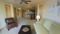 This single-floor unit offers an open-concept layout that seamlessly connects the living area to the kitchen, creating a spacious and functional flow. The living room is comfortably appointed with plush carpeted flooring, a ceiling fan with light fixture, and ample room for seating and a coffee table. Warm neutral tones on the walls and trim contribute to a welcoming ambiance throughout the space.

The kitchen is centrally positioned and features medium-toned wood cabinetry, dark countertops, a pendant light above the island or peninsula, and black appliances including a refrigerator. The kitchen peninsula provides additional prep space and serves as a natural divider between the living and cooking areas.

Visible from the living room, a dedicated laundry area with a washer is tucked conveniently within the unit, adding practical everyday functionality. Several interior doors lead to additional rooms, suggesting private spaces such as bedrooms or a bathroom, each framed with light wood trim that carries a consistent design language throughout.

Overall, the space presents a clean, practical, and cohesive interior with consistent warm neutral finishes, carpeted common areas, and an efficient open layout well-suited for comfortable day-to-day living.