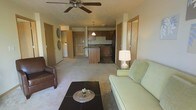 This single-floor unit offers an open-concept layout that seamlessly connects the living area to the kitchen, creating a spacious and functional flow. The living room is comfortably appointed with plush carpeted flooring, a ceiling fan with light fixture, and ample room for seating and a coffee table. Warm neutral tones on the walls and trim contribute to a welcoming ambiance throughout the space.

The kitchen is centrally positioned and features medium-toned wood cabinetry, dark countertops, a pendant light above the island or peninsula, and black appliances including a refrigerator. The kitchen peninsula provides additional prep space and serves as a natural divider between the living and cooking areas.

Visible from the living room, a dedicated laundry area with a washer is tucked conveniently within the unit, adding practical everyday functionality. Several interior doors lead to additional rooms, suggesting private spaces such as bedrooms or a bathroom, each framed with light wood trim that carries a consistent design language throughout.

Overall, the space presents a clean, practical, and cohesive interior with consistent warm neutral finishes, carpeted common areas, and an efficient open layout well-suited for comfortable day-to-day living.