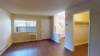 This single-story space showcases a straightforward, functional layout with warm hardwood-style flooring running throughout. The main living area is bright and open, benefiting from a large window that invites natural light and offers a view of the surrounding exterior. A wall-mounted air conditioning unit provides climate control directly in the living space. The textured ceiling adds a classic touch overhead, while neutral yellow-toned and gray walls create a clean, welcoming atmosphere. A white front door with a standard panel design serves as the main entry point. The living area flows seamlessly into the kitchen zone, which is defined by a half-wall counter that functions as a breakfast bar or pass-through surface. The kitchen area appears bright with lighter wall tones and continues the same hardwood-style flooring. Adjacent to the main living space, a doorway reveals a closet with built-in shelving, offering convenient storage. The overall layout is compact yet efficient, making practical use of every area. The space presents a clean, move-in-ready condition with simple, durable finishes throughout.
