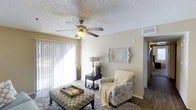 This single-floor residence offers a well-appointed and inviting living space designed for both comfort and functionality. The living room serves as the heart of the home, highlighted by large sliding glass doors dressed with vertical blinds that allow an abundance of natural light to fill the room. A ceiling fan with a built-in light fixture is centrally mounted on the textured ceiling, providing both airflow and ambient lighting. The space is thoughtfully furnished with a patterned accent chair, a plush area rug, and an upholstered ottoman that doubles as a coffee table, creating a cozy and functional seating arrangement. Neutral wall tones lend a warm, welcoming ambiance throughout, complemented by tasteful décor including a decorative mirror and accent greenery. Dark hardwood-style flooring lines the hallway leading further into the home, offering a rich contrast to the lighter living areas. The layout flows naturally from the main living space into additional areas of the residence, suggesting a practical and connected floor plan. Overall, the space strikes a balance between casual comfort and clean, understated style.