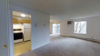 This single-floor residence offers a thoughtfully arranged layout with a spacious, open living area as its centerpiece. The living room is finished with wall-to-wall carpeting and benefits from abundant natural light, courtesy of a large sliding glass door that opens to an outdoor balcony and a separate window framing views of surrounding greenery. A wall-mounted air conditioning unit and a baseboard heater ensure year-round climate control.

The kitchen is set apart from the main living area, accessible through a dedicated doorway. It features tile flooring, wood-toned upper cabinetry, a full-size refrigerator, and a range, providing a functional and efficient cooking space.

The clean, neutral color palette throughout — with white walls and light-toned finishes — creates a bright and airy atmosphere that enhances the sense of openness. The combination of natural light from multiple windows, outdoor access, and a well-defined separation between living and kitchen areas makes this a practical and comfortable one-bedroom, one-bathroom residence.