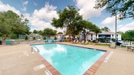 This two-floor residential community offers a well-appointed outdoor amenity area designed for both relaxation and recreation. At the heart of the outdoor space is a generously sized swimming pool with brick-tiled edging and clear blue water, complemented by a surrounding concrete sundeck furnished with lounge chairs and seating arrangements for residents to enjoy the open sky and natural surroundings.

Adjacent to the pool, a dedicated outdoor kitchen and grilling area features stone-clad counter structures equipped with stainless steel grills, providing an ideal setup for outdoor dining and social gatherings. The space is further enhanced by a tennis court with a bright blue surface, enclosed by a chain-link fence and visible from the pool area.

The entire amenity zone is enclosed by a wrought iron fence, adding a sense of security while maintaining an open, welcoming atmosphere. Mature trees throughout the property provide ample shade and contribute to a green, park-like setting. The residential buildings, finished in a white and blue color scheme, frame the outdoor space and reflect a clean, consistent architectural style. Overall, the community presents a functional and inviting outdoor environment suited to an active residential lifestyle.
