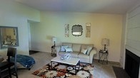 This single-floor space presents a thoughtfully arranged living room that combines comfort with decorative charm. The living area is finished with neutral, light-toned walls that create a calm and inviting atmosphere, complemented by wall-to-wall carpeting in a soft beige hue that extends throughout the space.

At the heart of the room, a cushioned sofa in a light gray upholstery serves as the focal point, dressed with accent throw pillows in muted teal tones. A rectangular white-topped coffee table anchors the seating area, accessorized with a spherical decorative piece that adds visual interest. Beneath the furniture arrangement sits a bold floral area rug featuring rich tones of brown, rust, and cream, providing a striking contrast to the otherwise neutral palette.

The walls are adorned with a curated arrangement of decorative mirrors — a central ornate framed mirror flanked by four smaller geometric square mirrors — adding depth and dimension to the space. Two matching black metal end tables on either side of the sofa each support a table lamp, providing balanced, warm ambient lighting throughout the room.
