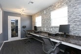This single-floor space is designed as a practical and polished workstation area, likely serving as a business center or administrative office. The room is finished with cool-toned, slate-blue walls that lend a calm and professional atmosphere throughout. Multiple windows fitted with white horizontal blinds allow ample natural light to filter into the space, creating a bright and welcoming environment.

A standout feature of this space is the elongated built-in desk finished with a rich, dark emperador marble-style countertop, providing generous workspace for multiple users simultaneously. The countertop runs the length of one wall and is accompanied by ergonomic task chairs, making it well-suited for productivity. Adjacent to the desk, a striking linear mosaic tile feature wall in earthy tones of brown, gray, and cream adds a refined decorative element and visual texture.

The space is equipped with multiple electrical outlets and data ports integrated neatly into the backsplash area, ensuring seamless connectivity for workstation setups. A glass-paneled interior door connects this room to an adjacent, brighter common area, promoting an open flow between spaces. Track lighting on the ceiling provides focused illumination across the workstations. The flooring consists of patterned carpet tiles, adding warmth and a professional finish to the overall design.