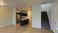 This two-story space is thoughtfully designed with a contemporary aesthetic that balances functionality and style. The main level showcases an open-concept kitchen that serves as a focal point, featuring rich dark cabinetry that contrasts elegantly with the light hardwood flooring extending throughout the space. A central kitchen island with a built-in sink provides additional workspace and acts as a natural gathering area. Stainless steel appliances, including a full-size refrigerator, microwave, and range, are seamlessly integrated into the cabinetry for a cohesive, modern look. Recessed lighting illuminates the kitchen evenly, creating a bright and functional environment. The open layout connects the kitchen to the adjacent living area, enhancing the sense of spaciousness on the main floor. Crisp white walls throughout contribute to an airy, clean atmosphere. A carpeted staircase visible from the main level provides access to the second floor, offering additional living space above. The overall design is clean and modern, with neutral tones and quality finishes that create a welcoming and versatile environment suitable for a variety of lifestyles.