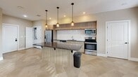 Located at 221 Trowbridge St in Grand Rapids, Michigan, this thoughtfully designed unit offers a single-floor layout that maximizes functionality within a compact footprint. The open-concept main area seamlessly integrates the living room, dining space, and kitchen, creating a bright and cohesive environment. Large windows line multiple walls, flooding the space with natural light and offering views of the surrounding greenery.

The kitchen is a standout feature, equipped with stainless steel appliances including a French door refrigerator, electric range, built-in microwave, and dishwasher. Contemporary light wood cabinetry extends to the ceiling, complemented by a white subway tile backsplash and dark granite countertops. A central island with matching granite provides additional prep space and casual seating, illuminated by a trio of stylish industrial pendant lights.

Throughout the unit, large-format herringbone-patterned tile flooring adds visual interest and continuity. A built-in Murphy bed cabinet in the main living area offers flexible sleeping arrangements. The bathroom is ADA-accessible, featuring a wall-mounted vanity with a wood base, grab bars, and matching tile flooring. Recessed lighting and neutral warm-toned walls complete the clean, modern aesthetic of this well-appointed space.