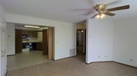 This single-story space offers a practical and functional layout centered around an open living area finished with wall-to-wall carpeting and neutral white walls. A ceiling fan with a built-in light fixture serves as a focal point overhead, providing both ventilation and ambient lighting to the main living space. A floor vent ensures efficient climate control throughout the area.

The living space connects directly to a kitchen, visible through a wide doorway. The kitchen features wood-toned cabinetry, open shelving, and under-cabinet fluorescent lighting, creating a warm and utilitarian cooking environment. Countertop workspace and additional storage are also present.

A hallway extends from the living area, leading to additional rooms within the unit. The corridor features track lighting overhead, hinting at further living or sleeping spaces beyond. The overall design is straightforward and efficient, with a neutral color palette of whites and beiges throughout, making the space feel clean and adaptable to various furnishing styles. The layout prioritizes connectivity between rooms, offering a cohesive and easy-to-navigate floor plan.