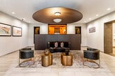 This single-floor lobby presents a carefully curated blend of contemporary design and upscale materials. The most striking architectural feature is a large, dark circular ceiling medallion, from which a pendant light is suspended, creating a bold focal point that anchors the entire space. Recessed lighting complements this centerpiece, casting a warm, even illumination throughout.

The flooring transitions between large-format, light-toned tiles and a richly patterned area rug that defines the central lounge seating area. This seating arrangement includes a curved black leather sofa accented with deep jewel-toned throw pillows, flanked by sleek barrel chairs with gold-toned metal bases. Cylindrical brass side tables complete the grouping, adding warmth and a luxurious touch.

A dark, studded partition wall serves as a visual divider, directing the eye toward a warm wood-paneled elevator vestibule behind it. Decorative wall niches featuring intricate lattice-style panels are integrated throughout, adding textural contrast against the smooth, neutral walls. Framed artwork is displayed thoughtfully along the walls.

A corridor extends toward a glazed entrance passage, where an exposed dark brick wall and large windows introduce natural light and an industrial edge. The overall ambiance is polished and welcoming, with a strong sense of refined, modern character.
