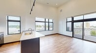 This contemporary single-floor unit showcases a thoughtfully designed open-concept layout with soaring ceilings that amplify the sense of space throughout. The living and dining area is bathed in natural light through oversized, dark-framed windows that frame sweeping urban views of the surrounding cityscape. Light-toned hardwood flooring runs throughout the main living spaces, complementing the clean, modern aesthetic.

The kitchen is a standout feature, anchored by a large island with a granite countertop and an undermount sink. Dark espresso cabinetry lines the walls, providing ample storage, while stainless steel appliances — including a refrigerator, electric range, and microwave — complete the space. Track lighting overhead adds functional illumination. The kitchen also benefits from multiple windows offering bright, open views.

A dedicated bedroom features plush carpeting, generous wall space, and access to a spacious walk-in closet equipped with wire shelving and hanging rods. The bathrooms maintain the same modern finishes, with dark wood vanities, granite countertops, and light hardwood-style flooring. A sliding glass door from the living area opens to a private balcony, offering elevated outdoor views of the surrounding neighborhood — a notable highlight of this well-appointed space.