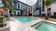 This outdoor amenity space is centered around a generously sized swimming pool with clear turquoise water, framed by a broad, stone-paver pool deck. Surrounding the pool, rows of orange-cushioned lounge chairs provide ample space for sunbathing, while the multi-story residential building wraps around the courtyard on multiple sides, creating an enclosed, resort-like atmosphere. Tall palm trees are positioned throughout the space, adding a tropical character and vertical visual interest against the building's bold white and blue exterior facade.

Adjacent to the main pool is a separate spa, finished with the same stone-paver decking for a cohesive aesthetic. A dedicated outdoor dining and lounge area features slatted tables paired with chairs, all shaded by large orange market umbrellas. Dense bamboo and tropical shrub plantings border this zone, creating a sense of privacy and a lush green backdrop.

A built-in outdoor grilling station is integrated into a landscaped garden area, surrounded by colorful tropical plantings including palms, variegated shrubs, and flowering plants with red mulch ground cover. Together, these features create a well-appointed, tropical outdoor retreat designed for relaxation and social gatherings.