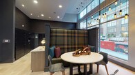 Located at 12 W Gay St in downtown Columbus, Ohio, this contemporary lobby space makes a striking first impression with its thoughtfully curated design and upscale finishes. The space is defined by soaring ceilings that amplify a sense of openness and grandeur, while floor-to-ceiling windows along the street-facing wall flood the interior with abundant natural light and offer views of the surrounding urban streetscape.

A standout feature is the bold, oversized banquette upholstered in a rich plaid fabric with warm gold, green, and charcoal tones, which serves as a dramatic focal point and inviting gathering area. Paired with a round marble-top table and eclectic accent chairs, the seating area blends classic and modern sensibilities seamlessly. Decorative wooden sculptural accents add an artisan touch to the tabletop styling.

Overhead, a curated arrangement of pendant lights in varied heights adds warmth and visual interest to the space. The lobby also incorporates a sleek reception or service counter finished in light wood tones, complemented by a wall-mounted mailbox or parcel area nearby. Dark, textured wall treatments throughout the space provide a sophisticated contrast to the light flooring, creating a polished and cohesive aesthetic that reflects the energy of its vibrant downtown Columbus location.