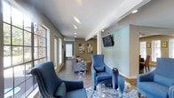 This single-floor communal space is thoughtfully designed to foster comfort and social interaction. The open-concept layout flows seamlessly from one area to the next, making the most of the available footprint. Recessed lighting throughout the ceiling provides consistent, well-distributed illumination across all zones.

The main lounge area features stylish blue upholstered armchairs with nail-head trim detailing, a neutral-toned sofa, and a glass-top coffee table with metallic accents, all arranged over a patterned blue area rug. Vibrant orange ottomans provide a bold pop of color, complementing the light blue accent wall on which a wall-mounted flat-screen television is prominently displayed.

Hardwood-style flooring runs throughout the space, lending a warm and cohesive aesthetic. Large windows along one side of the room allow natural light to filter in, enhancing the overall brightness of the interior. A secondary seating nook offers additional lounge chairs arranged for conversation.

Toward the rear of the space, a kitchenette area with rich wood cabinetry and a warm amber-orange accent wall provides a functional refreshment station. The overall design palette—combining cool blues, warm oranges, and neutral tones—creates an inviting and energetic ambiance suitable for a shared community or hospitality setting.