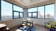 This contemporary upper-floor unit offers a bright and airy atmosphere, defined by expansive floor-to-ceiling windows with teal-framed accents that frame sweeping panoramic city views. Natural light pours throughout the space, highlighting the light wood-tone flooring and clean neutral walls.

The open-plan living and dining area is anchored by a coffered ceiling detail, lending architectural character to the space. Large corner windows maximize light and views, creating an inviting atmosphere for both relaxing and entertaining.

The kitchen is efficiently designed with sleek white cabinetry extending to the ceiling, stainless steel appliances including a full-size refrigerator, built-in microwave, electric cooktop, and dishwasher. A pendant light fixture adds a modern finishing touch overhead.

The bedroom is a sunlit retreat, benefiting from its own city-facing windows that continue the sweeping views found throughout the unit. The room accommodates a comfortable bed and offers a calm, restful ambiance.

The bathroom is clean and functional, featuring a single vanity with an integrated sink, a full mirror, and a tub-shower combination with ample storage. Wood-look flooring ties the bathroom seamlessly to the rest of the unit's design language.

Overall, the space presents a cohesive modern aesthetic with an emphasis on natural light, urban views, and functional living.