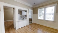 This single-floor space showcases a blend of classic architectural details and functional design. The main living area features warm, light-toned hardwood floors that extend throughout, creating a cohesive and inviting atmosphere. Elegant crown molding frames the ceilings, adding a refined, traditional character to the interior. Neutral taupe walls complement the crisp white trim and wainscoting, lending the space a clean and timeless aesthetic.

A standout feature is the pass-through kitchen, framed by a white paneled half-wall and accessible through a wide doorway. The kitchen is equipped with white cabinetry, a refrigerator, and a stove with a sink, and benefits from its own window that draws in natural light and provides views of the greenery outside. The pass-through counter adds both functionality and an open, connected feel between the kitchen and living areas.

Large windows in the main living space allow generous amounts of natural light to fill the room, enhancing the bright and airy ambiance. The wainscoting detail beneath the windows adds further architectural interest. Overall, the space offers a well-preserved, character-rich environment with thoughtful details that speak to its classic origins.