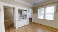 This single-floor space showcases a blend of classic architectural details and functional design. The main living area features warm, light-toned hardwood floors that extend throughout, creating a cohesive and inviting atmosphere. Elegant crown molding frames the ceilings, adding a refined, traditional character to the interior. Neutral taupe walls complement the crisp white trim and wainscoting, lending the space a clean and timeless aesthetic.

A standout feature is the pass-through kitchen, framed by a white paneled half-wall and accessible through a wide doorway. The kitchen is equipped with white cabinetry, a refrigerator, and a stove with a sink, and benefits from its own window that draws in natural light and provides views of the greenery outside. The pass-through counter adds both functionality and an open, connected feel between the kitchen and living areas.

Large windows in the main living space allow generous amounts of natural light to fill the room, enhancing the bright and airy ambiance. The wainscoting detail beneath the windows adds further architectural interest. Overall, the space offers a well-preserved, character-rich environment with thoughtful details that speak to its classic origins.