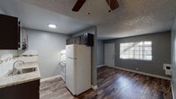 This single-floor unit showcases a thoughtfully updated interior with a cohesive modern aesthetic. The open living and kitchen area is unified by rich wood-look plank flooring and neutral gray walls, creating a clean, contemporary feel throughout. A ceiling fan with a pull-chain light fixture adds both function and a casual charm to the living space, which benefits from natural light streaming through a large window near the front entry door.

The kitchen is a standout feature, equipped with dark espresso cabinetry — including upper cabinets that extend to the ceiling for ample storage — granite-style countertops, a stainless steel sink, a glass tile backsplash with a decorative accent border, and a white electric range and refrigerator. The layout connects seamlessly to the living area, making the space feel open and functional.

The bedroom offers a generously sized, open layout with the same wood-look flooring, gray walls, and a textured ceiling. A wall-mounted heater unit and a window providing natural light are also present.

The bathroom is elegantly finished with large-format marble-look tile in the tub/shower surround, an accent mosaic tile border, a dark vanity with a white countertop and undermount sink, brushed nickel fixtures, and a well-lit vanity mirror. Tile flooring completes the polished look of this well-renovated space.