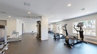 Located at 7288 Maple Street in Mission, BC, this dedicated fitness facility offers a spacious, single-floor layout designed to accommodate a comprehensive workout experience. The exercise room features a clean, modern aesthetic with white walls, smooth white ceilings, and durable dark rubber flooring throughout, providing both safety and functionality for a wide range of activities.

The space is generously equipped with a diverse array of fitness machines, including treadmills, ellipticals, stationary and spin bikes, cable and weight machines, and a leg press unit. A full dumbbell rack with kettlebells, along with built-in open shelving along one wall, adds convenience and organization to the facility.

Natural light fills the room through multiple windows and double-door access points on two sides, creating a bright and inviting atmosphere that enhances the overall workout environment. Recessed ceiling lighting ensures consistent illumination throughout. A water cooler is conveniently positioned near the exit, and the open floor plan allows ample space for free movement and stretching. The layout is thoughtfully organized, separating cardio equipment from strength training areas to maximize usability and flow throughout this well-appointed fitness space.