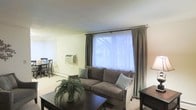 This single-floor space offers a welcoming and functional layout designed for comfortable everyday living. The living area serves as the heart of the home, centered around a plush, grey upholstered sofa accented with coordinating throw pillows. A dark wood coffee table anchors the seating arrangement, complemented by a matching side table that adds a sense of cohesion to the space. A large window dressed with elegant grey curtains and white blinds allows generous natural light to fill the room, creating a bright and airy atmosphere. A wall-mounted air conditioning unit provides climate control, adding to the practicality of the space. Adjacent to the living area, a dining space is visible, furnished with dark wood chairs and a matching table, offering a defined area for meals. The light-toned walls and carpeted flooring contribute to the overall neutral and calming palette throughout. A lush potted fern plant adds a touch of greenery and warmth, enhancing the inviting ambiance of the living space. The layout is thoughtfully arranged to maximize both comfort and functionality within a single floor plan.