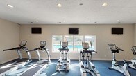 This fitness center is a purpose-built exercise facility designed to support a well-rounded cardio workout routine. The space is outfitted with multiple cardio machines, including treadmills and elliptical trainers, arranged to maximize usability and comfort. Durable blue fitness flooring covers the entire exercise area, providing cushioning and traction underfoot.

Natural light fills the room through a wide, multi-pane window along the far wall, which also offers a direct view of an outdoor swimming pool and surrounding recreational area — creating an energizing and visually open atmosphere during workouts. Two wall-mounted flat-screen televisions are positioned at opposite ends of the room, ensuring visibility from any machine and allowing users to stay entertained throughout their sessions.

Recessed lighting is evenly distributed across the textured ceiling, providing consistent, well-lit conditions throughout the day and evening. The neutral-toned walls keep the space feeling clean and focused. The single-floor layout ensures easy accessibility, and the overall design prioritizes functionality while maintaining a bright, welcoming environment. This fitness center serves as a convenient and practical amenity for those seeking on-site exercise facilities.