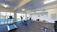 This single-floor fitness center offers a clean, functional layout designed to support a variety of workout routines. The space is divided into two distinct zones: a carpeted cardio area and a hard-floor stretching and flexibility zone. The cardio section is equipped with treadmills, elliptical trainers, and stationary bikes, providing ample options for aerobic exercise. The adjacent open area features stability balls and balance equipment, ideal for stretching and low-impact workouts.

Large floor-to-ceiling windows along one wall flood the interior with natural light and provide views of the surrounding outdoor environment, creating an open and energizing atmosphere. The ceiling is finished in white with a ceiling fan that aids in air circulation, complementing the recessed and fluorescent lighting fixtures that ensure consistent illumination throughout the space. The neutral white walls and two-toned flooring — dark carpet in the cardio zone and light hardwood-style flooring in the stretching area — contribute to a clean, organized aesthetic. Overall, the space is practical, bright, and well-maintained.