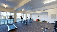 This single-floor fitness center offers a clean, functional layout designed to support a variety of workout routines. The space is divided into two distinct zones: a carpeted cardio area and a hard-floor stretching and flexibility zone. The cardio section is equipped with treadmills, elliptical trainers, and stationary bikes, providing ample options for aerobic exercise. The adjacent open area features stability balls and balance equipment, ideal for stretching and low-impact workouts.

Large floor-to-ceiling windows along one wall flood the interior with natural light and provide views of the surrounding outdoor environment, creating an open and energizing atmosphere. The ceiling is finished in white with a ceiling fan that aids in air circulation, complementing the recessed and fluorescent lighting fixtures that ensure consistent illumination throughout the space. The neutral white walls and two-toned flooring — dark carpet in the cardio zone and light hardwood-style flooring in the stretching area — contribute to a clean, organized aesthetic. Overall, the space is practical, bright, and well-maintained.
