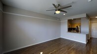 This single-story space in Richardson presents a clean, functional layout with modern finishes throughout. The main living area is defined by rich, dark hardwood flooring that contrasts elegantly with light gray walls and crisp white baseboards, creating a polished and inviting atmosphere. A stylish brushed-nickel ceiling fan with an integrated light fixture serves as a practical focal point overhead, ensuring comfortable airflow and ambient lighting throughout the room. Crown molding lines the upper perimeter of the walls, adding a refined architectural touch to the space. A notable feature is the pass-through kitchen window, which opens the living area to the kitchen beyond, enhancing connectivity and creating a sense of openness between the two spaces. The kitchen, visible through the pass-through, features warm cabinetry and what appears to be granite or stone countertops, lending a sophisticated feel. Recessed lighting in the kitchen adds to the overall brightness of the interior. The open, uncluttered layout of the living space offers flexibility for a variety of furniture arrangements, making it highly adaptable to different lifestyle needs. Overall, the space conveys a well-maintained, move-in-ready quality with thoughtful design details throughout.