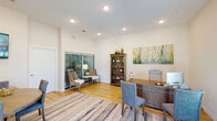 Located at 5010 Via Celeste in Pensacola, Florida, this single-story professional office space combines functionality with a polished, contemporary aesthetic. The interior is finished with warm hardwood-style flooring throughout, crisp white walls, and recessed lighting that ensures consistent, comfortable illumination across all areas.

The primary office area centers around a substantial wooden executive desk paired with an ergonomic chair and guest seating. A large nature-inspired canvas painting adorns the wall, contributing a calm, professional ambiance. A wooden bookcase provides both storage and decorative display, while a sliding glass door with blinds allows controlled natural light and access to an adjacent outdoor area. A seating nook near the sliding door offers a secondary conversation space with woven accent chairs.

A dedicated conference nook accommodates a round wooden table surrounded by upholstered chairs, a wall-mounted flat-screen television, and a decorative gold clock. This area is well-suited for small meetings or collaborative discussions.

Near the main entrance, double glass doors flood the space with natural light, complemented by a sideboard console and a decorative round mirror. A striped area rug anchors the open central zone, tying together the various functional areas into a cohesive, inviting professional environment.