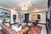 This single-floor amenity space is thoughtfully designed as a multifunctional social hub, blending recreational and lounge areas into a cohesive, well-appointed environment. The layout is divided into distinct zones, each with its own character while maintaining a unified aesthetic defined by polished concrete flooring, white walls, and bold dark trim and millwork throughout.

The games area anchors one side of the space, centered around a full-size black pool table set atop a gold patterned area rug, illuminated by a sleek cluster pendant light. Cue racks line the adjacent wall, and a counter-height bar table with leather chairs provides casual seating nearby. A large-format Scrabble board mounted on the wall adds a playful, oversized focal point, accompanied by a cozy lounge chair and side table.

The lounge zone features a tufted leather Chesterfield sofa, accent chairs, and a wood coffee table stacked with board games, all arranged before expansive floor-to-ceiling black-framed windows that frame direct views of an outdoor pool and courtyard, flooding the space with natural light.

A dining nook with booth-style seating sits adjacent to a deep green accent wall and oval mirror, while a decorative art-deco patterned wallcovering adds visual texture near a service counter. Together, these elements create an inviting, design-forward gathering space.