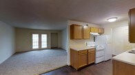 This single-floor space offers a practical and comfortable layout with an open-concept design that seamlessly connects the kitchen and living area. The kitchen is outfitted with warm honey-toned wooden cabinetry providing ample upper and lower storage, a white electric range with overhead ventilation, and a full-size refrigerator. Light-toned countertops complement the cabinetry, and the kitchen features durable dark hardwood-style flooring that adds contrast and visual depth. The adjacent living area transitions to soft, wall-to-wall carpeting, creating a cozy and inviting atmosphere. A standout feature of the living space is the set of classic French doors, which flood the room with natural light and offer a welcoming architectural detail. The textured ceiling and neutral wall tones throughout contribute to a clean, understated aesthetic. The overall layout is efficient and well-organized, making the most of the available space while maintaining a clear separation of function between the cooking and living zones. This unit is well-suited for those seeking a straightforward, move-in-ready space with essential amenities and a bright, open feel.