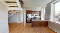 This distinctive two-floor space blends historic architectural character with modern finishes, creating a refined and dynamic living environment. The main level opens into a generous, light-filled living area with warm hardwood floors that extend throughout. A central staircase with a sleek cable-rail system connects the two floors, adding a contemporary industrial touch. The kitchen is well-appointed with rich dark wood cabinetry, stainless steel appliances including a French door refrigerator, an electric range, and a built-in microwave, all complemented by a large granite-topped island with an integrated sink. Track lighting illuminates the space efficiently. The upper level is a standout architectural feature, showcasing exposed white-painted timber roof trusses and skylights that draw in natural light. A cable-rail loft overlook connects to this dramatic upper space. Bedrooms on both levels offer hardwood floors and large windows with views of the surrounding historic urban streetscape. The bathrooms are elegantly finished — one features hexagonal mosaic tile flooring, a soaking tub, a glass-enclosed shower with wood-look tile surround, and a granite vanity, while a second bathroom offers large-format tile floors and a walk-in glass shower. The overall ambiance is that of a sophisticated urban loft with notable historic charm.