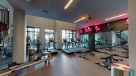 Located at 3600 E Fletcher Ave in Tampa, Florida, this two-story fitness center offers a comprehensive and well-equipped workout experience across multiple distinct zones. The main floor is anchored by a bright cardio area lined with treadmills, ellipticals, and a WaterRower, all positioned alongside large windows that invite natural light and outdoor views. A signature neon flamingo wall installation adds a bold, playful aesthetic to this section, complemented by sleek linear lighting and a dark ceiling treatment. Adjacent to the cardio zone is a strength training area featuring a full array of selectorized cable machines and weight equipment, set against floor-to-ceiling mirrors and accented with a teal color band. A dedicated training zone houses spin bikes, a tire flip station, heavy bag, battle ropes, and a comprehensive Training Zone rig, catering to high-intensity and functional fitness. The entry area features a dumbbell rack, adjustable bench, and wall-mounted display screen. A striking spiral staircase connects the floors, with the upper level offering additional strength equipment and open railings that overlook the surroundings. Throughout, rubber flooring, recessed lighting, and vibrant teal and magenta accent walls create an energetic and modern environment.