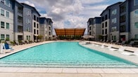 Located at 5745 Golden Triangle Blvd in Fort Worth, TX, this outdoor amenity space is designed to deliver a resort-style experience within a modern residential community. At the heart of the space is a generously sized, freeform swimming pool with crystal-clear turquoise water, surrounded by a broad concrete sun deck lined with contemporary white lounge chairs ideal for relaxing in the Texas sun. A shallow wading shelf along the pool's edge adds a leisurely, resort-inspired touch.

Flanking the pool on multiple sides are multi-story residential buildings clad in a combination of cool-toned blue and neutral gray siding, creating a cohesive and attractive architectural backdrop. Teal-accented market umbrellas provide additional shaded seating areas around the deck.

A large sail-style canopy shade structure anchors a dedicated lounge area, beneath which comfortable wicker seating with teal cushions surrounds a linear fireplace set into a rich, dark-tiled accent wall — perfect for evening gatherings. Adjacent to this is a built-in outdoor kitchen equipped with a stainless steel grill mounted on a handsome stone base, offering an ideal setup for outdoor entertaining. Natural stone and textured masonry wall detailing throughout the space add warmth and visual interest, complemented by lush greenery and mature trees along the perimeter.
