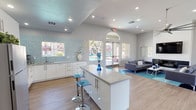 This single-floor space is designed with a clean, contemporary aesthetic that immediately conveys a welcoming and functional atmosphere. The open-plan layout seamlessly connects a central kitchenette area to a comfortable lounge, creating a versatile environment suited for gathering or hosting. 

At the heart of the space sits a sleek white island with a smooth countertop surface, accompanied by modern acrylic bar stools in a cool blue tone. The kitchenette features white cabinetry with a mosaic tile backsplash in aqua and white, adding a vibrant yet refined decorative accent. Recessed lighting throughout the ceiling ensures the space is well-lit, while vaulted ceiling lines add a sense of vertical openness.

The lounge area is furnished with contemporary grey sofas set atop a teal area rug, creating a cohesive color palette that ties the space together. Large glass doors with arched transom detailing draw the eye toward the exterior, offering direct views of an outdoor pool and allowing abundant natural light to flood the interior. Light-toned wood-style flooring runs throughout, enhancing the airy and modern feel. Overall, the space exudes a bright, polished, and inviting ambiance with thoughtful design details throughout.