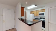 This renovated one-bedroom, one-bathroom unit offers a well-designed single-floor layout that maximizes functionality and comfort. The kitchen serves as a central highlight, featuring warm honey-toned wood cabinetry that provides ample storage space. A peninsula with a dark granite-style countertop creates a natural division between the kitchen and living areas, offering additional workspace and a casual dining or seating opportunity. The kitchen is equipped with a full suite of appliances, including a refrigerator, electric range, built-in microwave, and additional counter space for meal preparation. Recessed and flush-mounted ceiling lighting ensures the kitchen remains well-lit throughout the day. The light-toned tile flooring adds a clean, neutral aesthetic that complements the cabinetry and countertops. White walls and bright lighting contribute to an open, airy feel throughout the space. A hallway visible from the kitchen suggests convenient access to additional living areas, including the bedroom and bathroom, maintaining a practical and efficient floor plan overall.