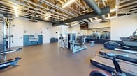 Located at 621 Suhtai Ct in Virginia Beach, Virginia, this single-floor fitness facility showcases a thoughtfully designed interior that blends industrial aesthetics with functional workout spaces. The exposed roof trusses and open ceiling structure create a sense of generous vertical space, while large black HVAC ducts run prominently overhead, contributing to the facility's industrial character. Suspended linear lighting fixtures illuminate the space effectively throughout.

The workout floor is finished with durable, neutral-toned concrete or rubberized flooring suited for high-traffic fitness use. Full-length mirrors line multiple walls, visually expanding the space and providing users with proper form-checking capability. The facility is divided into distinct training zones: a cardio area features treadmills, stationary bikes, and other aerobic machines positioned near large windows that invite natural light and outdoor views, while a strength training zone houses a comprehensive selection of resistance machines, cable systems, a squat rack, and a full dumbbell rack.

The walls feature a two-tone treatment with a dark navy lower panel and light upper walls. A water fountain and additional utility amenities are integrated along one wall. Louvered doors lead to additional rooms, and multiple access points throughout the space ensure smooth circulation. The overall ambiance is clean, modern, and purpose-built for fitness.
