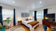 Located at 990 Palm Ave in the heart of West Hollywood, CA, this thoughtfully designed single-floor studio blends mid-century modern aesthetics with contemporary comfort. Warm hardwood flooring flows throughout the open layout, connecting the living, sleeping, and working areas seamlessly.

The main living space features a rich emerald green velvet sofa accented with olive and cream cushions, paired with a wooden desk and a blue ergonomic chair — ideal for both relaxing and working from home. A round arched mirror adds a stylish touch near the entry.

The bedroom area is a tranquil retreat, highlighted by large windows dressed in deep navy curtains that offer both ambiance and light control. A triptych of bold black-and-white geometric artwork anchors the space above the bed, flanked by warm-toned nightstands with gold sconce lighting. Recessed ceiling lights and light hardwood floors complete the inviting atmosphere.

The compact kitchen is efficiently designed with cream cabinetry, open shelving, a gas range, stainless steel appliances, and a built-in dark wood bar counter with industrial-style stools — perfect for casual dining.

The bathroom features clean white subway tile surrounding a soaking tub and shower combination, with brushed brass fixtures and a frosted window for natural light and privacy.