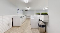 Located at 25 Fisherville Rd in Toronto, this shared laundry facility is a well-organized, single-floor amenity space designed for practical, everyday use. The layout comprises two dedicated laundry rooms, a connecting hallway, and a separate waiting area, all finished with a consistent, clean aesthetic throughout.

The primary laundry room is generously sized and equipped with multiple rows of white top-load washing machines and dryers, providing ample capacity for building residents. White subway tile walls, speckled terrazzo flooring, and bright fluorescent lighting create a clean, functional environment. A long wooden folding counter runs along one wall, offering convenient space for sorting and folding laundry. Potted greenery along the top of the machine banks adds a subtle touch of freshness to the utilitarian space.

The second laundry area features additional machines alongside white tiled walls, maintaining the same cohesive design language. The connecting hallway, finished with matching terrazzo floors and tiled walls, leads between the rooms and provides access to the facility's entry points, with posted informational notices for residents.

A separate waiting area offers a welcoming respite, distinguished by light wood-look flooring, glossy white horizontal tile walls, and a wall-mounted open shelf unit. A comfortable armchair and a compact side ottoman furnish the space, making it a practical spot to wait during laundry cycles.