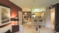 This single-floor residence showcases an open-concept layout that seamlessly connects the living, dining, and kitchen areas into one cohesive living environment. The kitchen is positioned behind a half-wall peninsula, creating a functional breakfast bar with sleek bar stools — ideal for casual dining or entertaining. Light wood cabinetry, a white refrigerator, and a pass-through design keep the kitchen feeling bright and accessible. Crown molding runs throughout, adding a refined architectural detail to the ceilings. A bold, deep red accent wall anchors the living area, complementing the neutral tile and carpeted flooring that transitions between zones. The dining area, visible through the open layout, is illuminated by a contemporary pendant light and features modern seating. Warm ambient lighting from table lamps and overhead fixtures creates an inviting atmosphere throughout. A ceiling fan contributes to the comfortable, well-ventilated feel of the space. The overall aesthetic balances contemporary design with warm, rich tones, resulting in a welcoming and functional single-level living environment.
