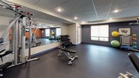 This fitness center is a comprehensively outfitted single-floor exercise facility designed to support a full range of workout routines. The space is organized into distinct functional zones, each catering to different training needs. A dedicated cardio area features treadmills, an elliptical trainer, and a recumbent stationary bike, all positioned in front of wall-mounted flat-screen televisions set against a bold orange accent wall, creating an energizing atmosphere for endurance training.

The strength-training section includes a multi-function cable machine, a leg press and resistance machines, and a weight bench paired with a tiered dumbbell rack holding a varied selection of free weights. Exercise balls and additional accessories are stored nearby for flexibility and functional training. A large floor-to-ceiling mirror spans one wall, enhancing the sense of openness and allowing users to monitor their form.

Throughout the space, durable rubber flooring provides traction and cushioning underfoot. Recessed lighting embedded in a drop-tile ceiling delivers consistent, even illumination. Roller-shade windows allow for natural light while maintaining privacy. The overall ambiance is clean, practical, and well-maintained, making it a functional environment suited for a broad spectrum of fitness activities.