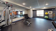 This fitness center is a comprehensively outfitted single-floor exercise facility designed to support a full range of workout routines. The space is organized into distinct functional zones, each catering to different training needs. A dedicated cardio area features treadmills, an elliptical trainer, and a recumbent stationary bike, all positioned in front of wall-mounted flat-screen televisions set against a bold orange accent wall, creating an energizing atmosphere for endurance training.

The strength-training section includes a multi-function cable machine, a leg press and resistance machines, and a weight bench paired with a tiered dumbbell rack holding a varied selection of free weights. Exercise balls and additional accessories are stored nearby for flexibility and functional training. A large floor-to-ceiling mirror spans one wall, enhancing the sense of openness and allowing users to monitor their form.

Throughout the space, durable rubber flooring provides traction and cushioning underfoot. Recessed lighting embedded in a drop-tile ceiling delivers consistent, even illumination. Roller-shade windows allow for natural light while maintaining privacy. The overall ambiance is clean, practical, and well-maintained, making it a functional environment suited for a broad spectrum of fitness activities.
