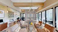 This thoughtfully designed single-floor amenity space blends comfort and functionality across a generously sized open-plan layout. The lounge area is anchored by soaring ceilings and floor-to-ceiling windows that flood the room with abundant natural light, framing outdoor views of lush landscaping. Large-format tile flooring and crisp white walls create a clean, airy backdrop throughout.

A striking metallic lattice room divider serves as a standout architectural element, elegantly separating the lounge seating area from the coworking zone without fully enclosing either space. Warm wood ceiling beams and accent panels add texture and contrast, complementing the contemporary pendant lighting fixtures that hang at varying heights throughout.

The lounge is furnished with plush sofas and accent chairs arranged around stylish coffee tables atop a richly patterned area rug. Adjacent to the lounge, a long white island bar with bar-height seating is flanked by a built-in refreshment station with wood cabinetry and a wall-mounted television framed by a dramatic slatted wood feature wall.

The coworking area provides multiple workstations with desktop computers and ergonomic seating, as well as a conference-style table for collaborative use. Accent walls with soft watercolor-effect murals and curated artwork add a sophisticated, gallery-like quality to the overall space.