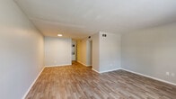 This single-floor unit offers a clean, modern aesthetic throughout its thoughtfully designed layout. The open living area is finished with durable wood-look plank flooring in a warm neutral tone, complemented by soft gray walls and crisp white baseboards. Recessed lighting and ceiling-mounted vents contribute to a tidy, well-maintained interior. The space flows naturally toward a galley-style kitchen, which features white shaker cabinetry with brushed metal hardware, granite-look countertops, a classic white subway tile backsplash, and black appliances including a refrigerator, range, and over-the-range microwave. A window on the exterior door allows additional natural light into the kitchen. The bedrooms are carpeted in a soft gray tone and each benefit from windows fitted with horizontal blinds, inviting views of surrounding greenery. The bathroom presents a clean, functional design with wood-look tile flooring, a floating white vanity, chrome fixtures, and a full tub-shower combination enclosed in white tile. The overall layout, as seen in the dollhouse view, is efficient and well-proportioned, with clearly defined zones for living, cooking, sleeping, and bathing — making this a practical and attractive single-level space.