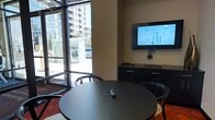 This single-floor space functions as a contemporary meeting or conference room, thoughtfully designed to balance functionality with a bright, professional atmosphere. Large floor-to-ceiling windows dominate one wall, flooding the interior with abundant natural light and offering open views of the surrounding urban streetscape, including neighboring high-rise buildings and outdoor activity. This generous glazing creates a strong visual connection to the outside while lending the room an airy, open feel. The meeting area centers around a round, dark-toned table paired with sleek matching chairs, fostering an inclusive, collaborative seating arrangement well-suited for small group discussions or presentations. A wall-mounted flat-screen display positioned on a neutral-toned accent wall serves as the room's primary presentation focal point, accompanied by a low-profile dark credenza below that provides additional storage and surface space for media equipment. The flooring features a bold, patterned area rug that adds a layer of visual warmth and definition to the meeting zone. Overall, the space conveys a polished, professional ambiance with clean lines, a restrained color palette of dark furnishings against light walls, and an emphasis on natural light — making it a highly functional environment for meetings, presentations, or collaborative work sessions.