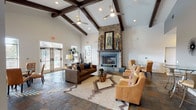 Located at 8711 Bowens Crossing St in San Antonio, Texas, this single-story community clubhouse delivers an impressive blend of rustic charm and modern sophistication. The centerpiece of the main lounge is a dramatic floor-to-ceiling stacked stone fireplace, flanked by vaulted ceilings adorned with dark wood exposed beams and dual ceiling fans, creating an airy, grand atmosphere throughout the space. Rich multicolored slate tile flooring flows seamlessly across the main areas, grounding the warm, inviting aesthetic. The lounge is thoughtfully arranged with leather seating, plush armchairs, and casual dining tables positioned near large windows that invite abundant natural light and outdoor views. A bright and welcoming entryway area features elegant French doors, tall windows dressed with sheer white curtains, and tasteful seating nooks accented by indoor greenery. Adjacent to the main lounge, a business center and media area offer built-in white cabinetry, a mounted flat-screen television, and dedicated workstations. A separate conference room provides a more focused environment, complete with a substantial wooden table, upholstered chairs, a statement chandelier, and coordinating built-in shelving with computer stations—ideal for meetings or collaborative work.