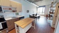 Located at 624 Yale Ave N in Seattle's vibrant South Lake Union neighborhood, this single-floor unit blends modern design with industrial character. The open-concept layout seamlessly connects the kitchen, dining, and living areas, creating a spacious and functional flow throughout.

The kitchen is outfitted with warm two-tone cabinetry — light wood uppers paired with darker lower cabinets — and features a stainless steel sink, dishwasher, and a freestanding butcher block island that provides additional prep space and storage. The kitchen opens directly to the dining area, which comfortably accommodates a full dining table and chairs, making it well-suited for both everyday living and entertaining.

The living area is bathed in natural light from floor-to-ceiling windows that frame outdoor greenery views, creating a bright and inviting atmosphere. Rich hardwood flooring runs throughout the space, adding warmth that contrasts beautifully with the exposed concrete ceiling and industrial-style ductwork above — hallmark features of the building's loft-inspired aesthetic. Polished pendant lighting complements the space's modern sensibility. Overall, the unit offers a refined urban living experience with thoughtful finishes and an airy, open ambiance.