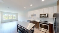 This contemporary single-floor unit showcases a clean, modern design with light wood-tone flooring and a neutral color palette that runs seamlessly throughout. The open-concept layout connects the kitchen and living area, creating a bright and functional living space filled with natural light from large windows overlooking lush greenery.

The kitchen is a standout feature, outfitted with a two-tone cabinetry scheme pairing crisp white upper cabinets with rich dark wood lower cabinets. A white quartz countertop and a classic subway tile backsplash add refinement, while a full suite of stainless steel appliances — including a French door refrigerator, range, over-the-range microwave, and dishwasher — provides a polished, move-in-ready finish. A pull-down spray faucet over the undermount sink adds practical elegance. A freestanding stainless steel kitchen island offers additional prep and storage space.

The bedroom is a serene, well-proportioned room bathed in natural light through wide windows with pleasant outdoor views. An adjacent closet provides convenient storage. The bathroom features a dark wood vanity with a white countertop, a framed mirror, and neutral tile flooring, maintaining the unit's cohesive contemporary aesthetic. Recessed lighting throughout ensures well-distributed illumination across all areas.