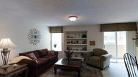 This single-floor residence offers a comfortable and thoughtfully arranged living space with a classic, homey aesthetic. The living room serves as the heart of the home, featuring a built-in shelving unit that provides both display and storage space, showcasing decorative accents and books. The room is anchored by a plush sofa and a coordinating upholstered armchair, arranged around a dark wood coffee table atop a patterned area rug, creating an inviting conversational setting. Multiple windows dressed with valances allow natural light to brighten the space, contributing to an open and airy feel. A textured popcorn ceiling adds a traditional character to the room, while a flush-mount ceiling light fixture provides warm, ambient illumination. Decorative wall art, including an ornate metal wall piece, adds a personalized touch to the neutral-toned walls. The overall ambiance is warm and welcoming, with a traditional design sensibility that emphasizes comfort and functionality. The layout is efficiently organized, making the most of the available floor space while maintaining a relaxed, livable atmosphere throughout.