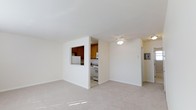 This single-floor residence offers a clean, functional layout designed for comfortable everyday living. The spacious living area is finished with soft wall-to-wall carpeting, creating a warm and inviting atmosphere. Crisp white walls and a ceiling fan with a light fixture contribute to a bright, airy feel throughout the main living space.

The kitchen is accessible via a dedicated entryway and is also connected to the living area through a convenient pass-through window — a practical design detail that enhances openness and communication between spaces. The kitchen features warm-toned wood cabinetry that provides ample storage, complemented by white appliances including a refrigerator, range, and microwave. The kitchen floor transitions to a light-toned hard surface, offering easy maintenance in this high-use area.

A refrigerator with an in-door dispenser is also visible along a side wall, adding to the functional amenities of the space. The overall design is straightforward and practical, with a neutral color palette that lends itself well to a variety of furnishing styles. The layout flows naturally between the living and kitchen areas, making the space feel cohesive and well-organized throughout.
