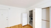 This single-floor unit offers a straightforward and functional layout that makes efficient use of its space. The living area is the largest zone in the unit, featuring warm hardwood flooring that lends a natural, inviting feel to the room. Large windows allow natural light to fill the space, creating a bright and airy atmosphere throughout. The white walls throughout the unit contribute to a clean, neutral aesthetic that provides a versatile backdrop for any style of furnishing.

The kitchen is a separate, dedicated space accessed directly from the living area and is finished with classic white tile flooring. It is equipped with a gas range and oven, a full-size top-freezer refrigerator, a sink with cabinetry, and additional upper and lower cabinet storage, providing ample room for everyday cooking needs. The kitchen's compact, efficient design keeps all essentials within easy reach.

A bathroom is also included within the unit, positioned adjacent to the kitchen area. The overall layout of the unit is well-organized, with each functional zone clearly defined while maintaining a sense of connectivity between spaces. The consistent use of white finishes and natural wood tones throughout creates a cohesive and tidy living environment across the entire floor plan.
